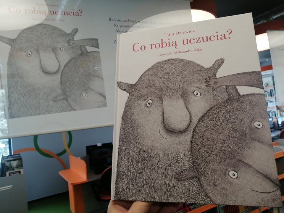 Na środku widać książkę "Co robią uczucia?", na okładce sympatyczne puchate stworki (uosabiające uczucia), w tle plansze wystawy oraz sala biblioteczna. 