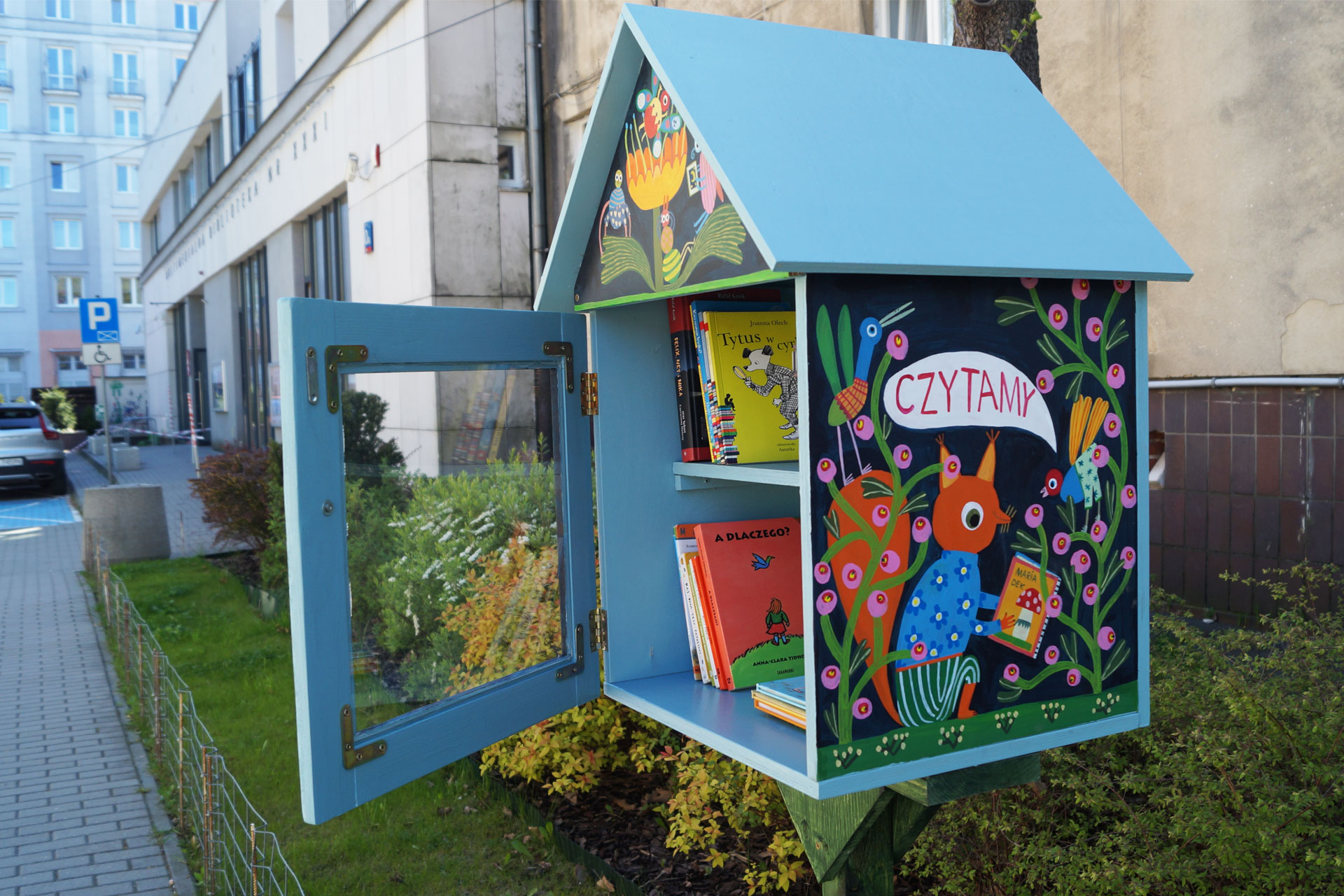 Kolorowy, drewniany domek na książki postawiony na nóżce, na świeżym powietrzu. Na ściance domku malunek wiewiórki, która czyta książkę. W tle budynek Multimedialnej Biblioteki. 